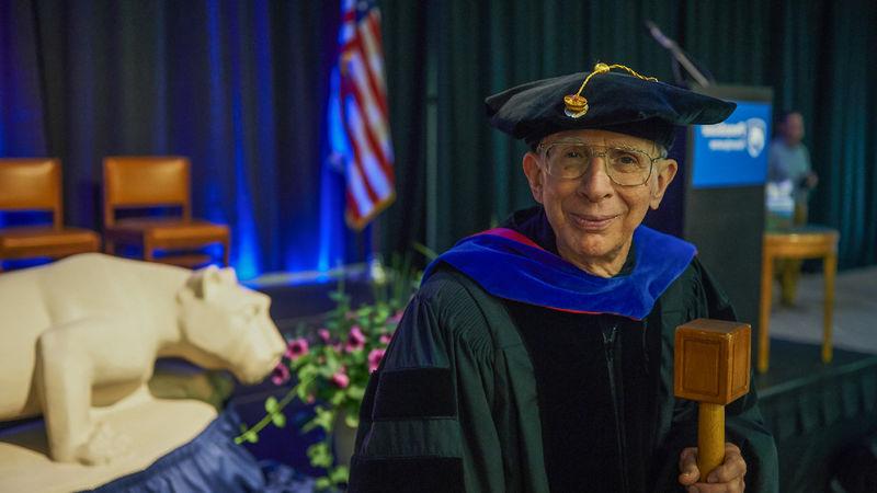 A man wears academic regalia and holds a mace.